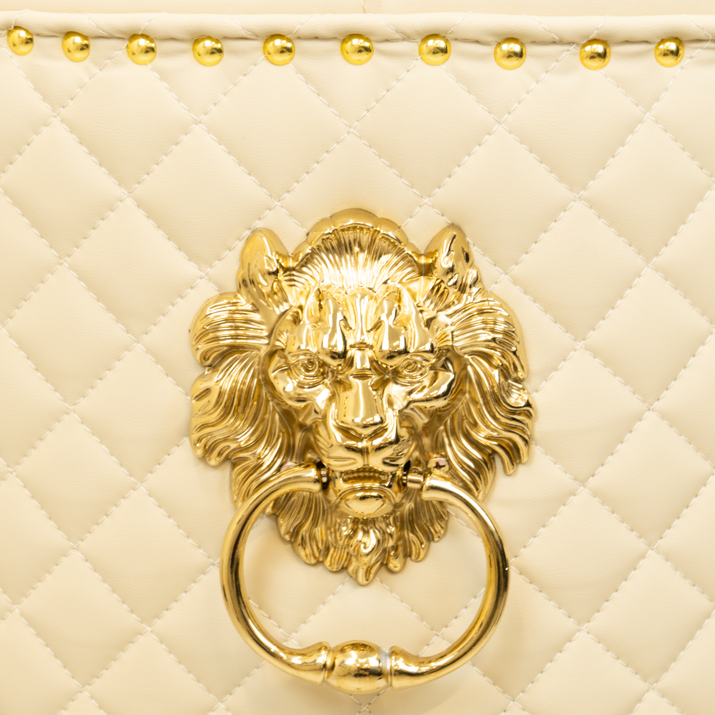 Close-up of a gold lion head ring pull mounted on a beige diamond-quilted upholstered chair back, with gold nailhead trim visible along the top edge.