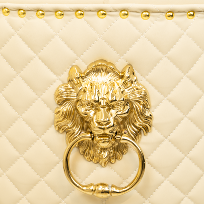 Close-up of a gold lion head ring pull mounted on a beige diamond-quilted upholstered chair back, with gold nailhead trim visible along the top edge.